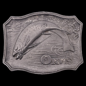 May include: A silver-colored metal belt buckle with a detailed relief of a fish leaping out of water, with a fishing line and the word "ORVIS" below. The buckle has a rectangular shape with a decorative border.