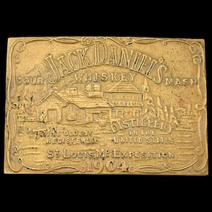 May include: A rectangular, gold-colored metal plaque with the words "Jack Daniel's Whiskey Mash" at the top. The plaque features an embossed image of a distillery and the text "The Oldest Registered Distillery on the United States, St. Louis Mo. Exposition 1904."