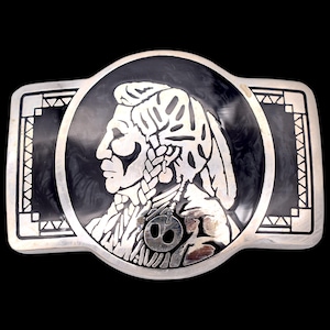 May include: Silver-toned belt buckle featuring a Native American profile in a circular design. The buckle has a black background and a decorative geometric border. The Native American figure is detailed with intricate silver patterns.