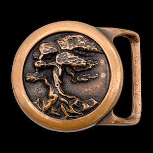 May include: A bronze-colored belt buckle with a detailed relief of a bonsai tree. The tree is centered within a circular frame, and the buckle has a rectangular bar for the belt. The design is set against a dark background.
