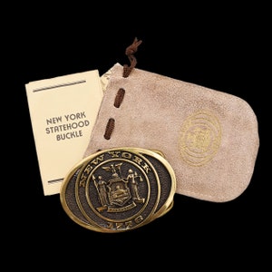 May include: Oval brass belt buckle with the words "New York" and "1776" engraved. The buckle features the New York state seal and comes with a tan suede pouch and a card that reads "New York Statehood Buckle."