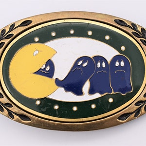 May include: A brass belt buckle with a Pac-Man design. The buckle features a yellow Pac-Man eating three blue ghosts on a white background. The background is surrounded by a green and gold border.