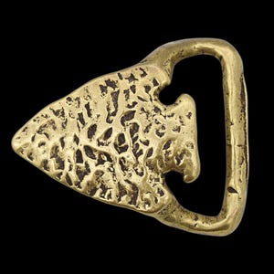 May include: A vintage brass belt buckle with an arrowhead design. The buckle has a textured surface and a D-shaped loop. The arrowhead has a cutout detail near the loop. The buckle is set against a black background.