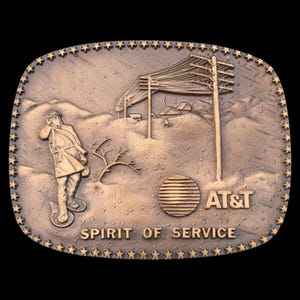 May include: Bronze-colored belt buckle with a relief design. It features a figure in a coat, telephone poles, and the text "AT&T SPIRIT OF SERVICE." The buckle has a star-studded border and a rectangular shape.