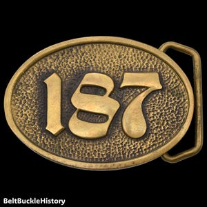 May include: An oval brass belt buckle featuring a textured, hammered background and the raised, polished number 187 centered on the face. The buckle includes a rectangular loop on the right side for attachment to a belt strap.