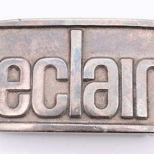 May include: A rectangular silver-colored belt buckle with the word "eclair" in bold, raised letters. The buckle has a slightly textured surface and a simple, classic design. The letters are in a sans-serif font.
