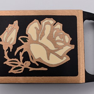 May include: A rectangular belt buckle with a rose design. The buckle features a black background with a cream-colored rose and green accents, framed by a copper-colored border. The handle is black.