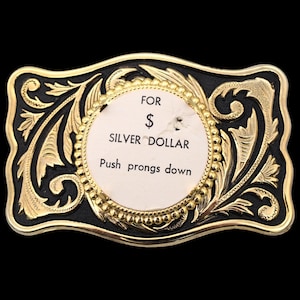 May include: A rectangular belt buckle with a black background and gold filigree design. The center features a circular area with the text "FOR $ SILVER DOLLAR Push prongs down."