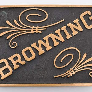 May include: A black metal belt buckle with the word "BROWNING" in gold lettering. The buckle has two gold stylized floral designs on either side of the word.