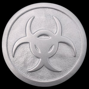 May include: A silver-colored metal disc with a raised biohazard symbol in the center. The symbol is a stylized three-pronged design with a circle in the center.