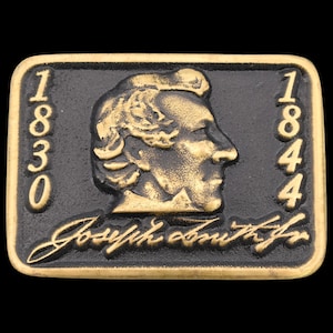 May include: Rectangular, two-tone metal belt buckle. The buckle features a profile of a man's face in gold against a black background. The dates 1830 and 1844 are on either side of the profile. The name "Joseph Smith Jr." is written in cursive.