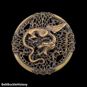 May include: A circular brass belt buckle featuring an intricate openwork web design. Centered on the lattice is a raised, detailed figure of a winged dragon with a long, curved tail. The metal has a polished gold-tone finish with dark patina.