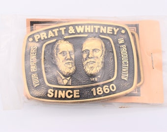 May include: A rectangular brass belt buckle featuring portraits of two men. The buckle is inscribed with "PRATT & WHITNEY" and "YOUR PARTNERS IN PRODUCTIVITY SINCE 1860". The buckle is in a clear plastic package.