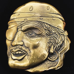 May include: A gold-toned metal belt buckle featuring a detailed pirate head with an eyepatch, a bandana, and a beard.