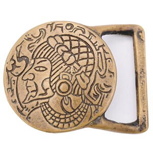 May include: A bronze-colored belt buckle with an engraved design of a profile of a person with elaborate headdress and surrounding glyphs. The buckle has a rectangular loop on one side.