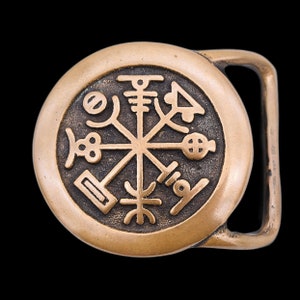 May include: A bronze-colored belt buckle with a circular design featuring an intricate, stylized compass rose symbol. The buckle has a rectangular bar for the belt strap and is set against a black background.
