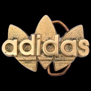 May include: A vintage gold-colored belt buckle featuring the Adidas logo. The buckle has a textured surface and a copper-colored loop for the belt. The Adidas text is in a bold, sans-serif font.