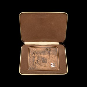 May include: A vintage copper-toned metal belt buckle with a relief scene of a person on a unicycle, telephone poles, and a small emblem. The buckle is presented in a brown velvet-lined box with a gold-colored trim.