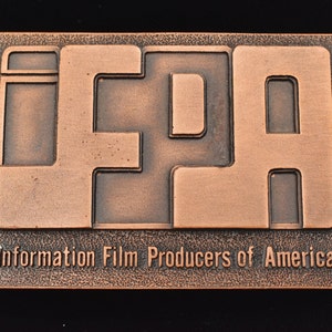 May include: A copper-colored belt buckle with the letters "IFPA" in a blocky font. The words "Information Film Producers of America" are engraved below the letters. The buckle has a rectangular shape and a textured border.