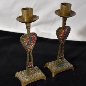 May include: A pair of brass candle holders with a heart-shaped design. The hearts are decorated with colorful enamel and the words "To the Lord our God" and "Us He Saves".