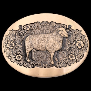 May include: Oval-shaped, bronze-colored belt buckle featuring a detailed relief of a sheep. The sheep is surrounded by floral and scrollwork designs. The buckle has a polished finish and is set against a black background.