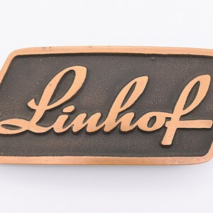 May include: A rectangular bronze-colored metal emblem with the word "Linhof" in a cursive font. The lettering is raised and in a lighter bronze color than the background. The emblem has a slightly beveled edge.