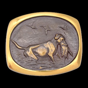May include: A rectangular belt buckle with a gold-colored border. The center features a bronze-colored relief of a dog carrying a bird in its mouth, with ducks flying in the background. The dog is depicted in water.