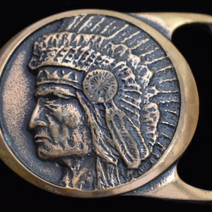 May include: A vintage belt buckle with a detailed relief of a Native American chief in profile. The buckle is circular with a bronze-colored chief and a gold-colored frame and clasp. The design features a feathered headdress.