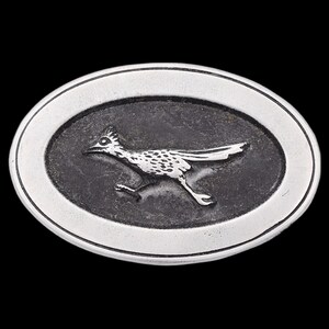 May include: Oval-shaped silver belt buckle featuring a detailed roadrunner design. The roadrunner is depicted in a running pose, with its body covered in speckled patterns. The buckle has a dark gray background and a silver border.