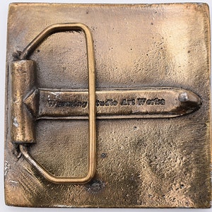 K&ouml;nnte beinhalten: Eine quadratische G&uuml;rtelschnalle aus Messing mit glatter Oberfl&auml;che. Die Schnalle hat eine Metallschaufel und eine Zunge. Die Schnalle ist mit dem Text "Wyoming Studio Art Works" graviert.