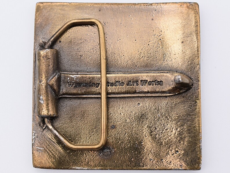 K&ouml;nnte beinhalten: Eine quadratische G&uuml;rtelschnalle aus Messing mit glatter Oberfl&auml;che. Die Schnalle hat eine Metallschaufel und eine Zunge. Die Schnalle ist mit dem Text "Wyoming Studio Art Works" graviert.