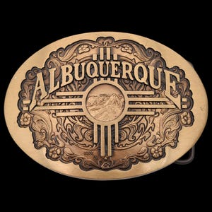 May include: Oval-shaped brass belt buckle with the word "ALBUQUERQUE" in raised lettering. The buckle features a central design with a mountain scene and a stylized sun symbol, surrounded by floral and scrollwork details.