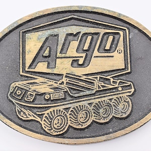 May include: Oval-shaped belt buckle with a black background and a gold-colored design. The design features the word "ARGO" in a gold box, and a detailed illustration of an amphibious vehicle.
