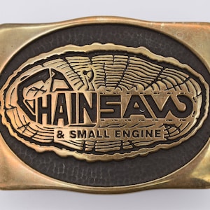 May include: A gold-colored metal belt buckle with a black oval inset. The oval features a stylized tree ring design with the text "CHAINSAWS & SMALL ENGINE" in black lettering.