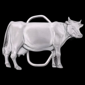 May include: A silver-colored metal belt buckle in the shape of a cow. The cow is standing, with detailed features including horns, udder, and a curved metal bar for the belt loop. The background is black.