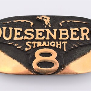 May include: A black and copper-colored metal belt buckle with an eagle design and the text "Duesenberg Straight 8".