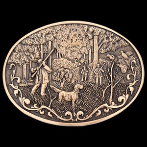 May include: Oval-shaped, bronze-colored belt buckle with a hunting scene. The detailed relief depicts hunters, a dog, trees, and birds in flight. Decorative scrollwork borders the scene, set against a black background.