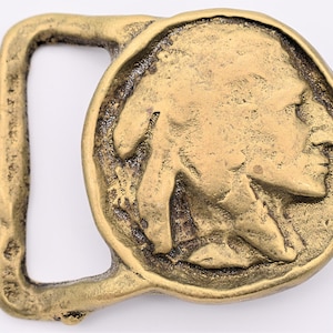 May include: A vintage brass belt buckle featuring a detailed relief of a Native American profile within a circular frame. The buckle has a rectangular loop and a textured, aged appearance.