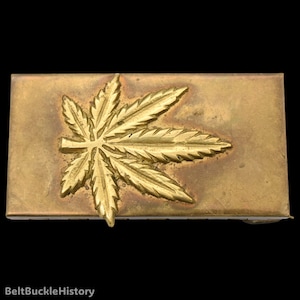 May include: A rectangular brass belt buckle featuring a raised, embossed cannabis leaf design centered on the front surface. The metal has a weathered, aged patina with a golden-brown tone and visible texture across the flat rectangular plate.