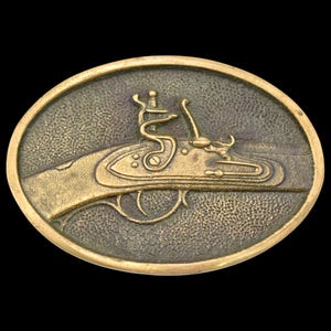 May include: Oval-shaped, antique-style belt buckle with a detailed relief of a flintlock pistol. The buckle has a bronze finish with a textured surface, set against a black background. The pistol is depicted in profile, showcasing its intricate design.