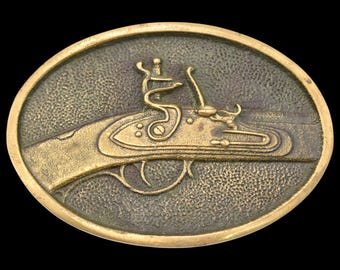 May include: Oval-shaped, antique-style belt buckle with a detailed relief of a flintlock pistol. The buckle has a bronze finish with a textured surface, set against a black background. The pistol is depicted in profile, showcasing its intricate design.