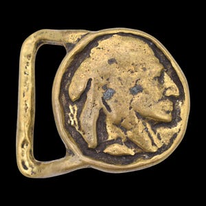 May include: A vintage brass belt buckle featuring a detailed relief of a Native American profile within a circular frame. The buckle has a rectangular loop and a tarnished, aged appearance, suggesting it is an antique or a vintage piece. The background is black.