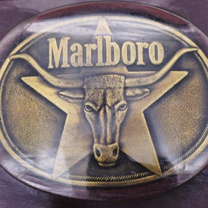 May include: Oval-shaped belt buckle with a gold-colored bull's head and star design. The word "Marlboro" is displayed in gold lettering across the top. The buckle has a dark brown border.