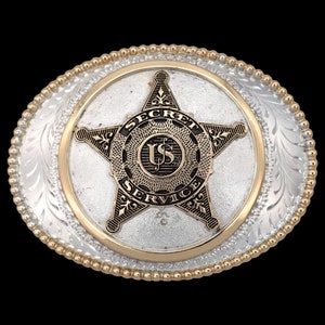 May include: Oval-shaped silver and gold belt buckle with a detailed design. The center features a star with the words "SECRET SERVICE" and "US" in black. The buckle has a beaded gold edge and a floral pattern.