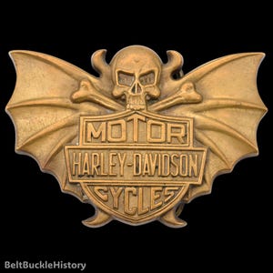 May include: A brass-toned metal belt buckle featuring a central shield with the embossed text MOTOR HARLEY-DAVIDSON CYCLES. The design includes a skull positioned above the shield, flanked by two large, stylized bat-like wings extending outward.