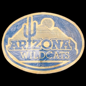 May include: An oval-shaped Arizona Wildcats belt buckle. The buckle features a blue background with gold lettering and a gold outline. The design includes a saguaro cactus and mountain range. The text "ARIZONA WILDCATS" is prominently displayed.