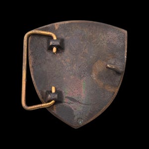 May include: A vintage belt buckle with a shield shape. The buckle is made of a dark, aged metal with a gold-colored bar and attachment points. The surface shows signs of wear and patina, suggesting age.