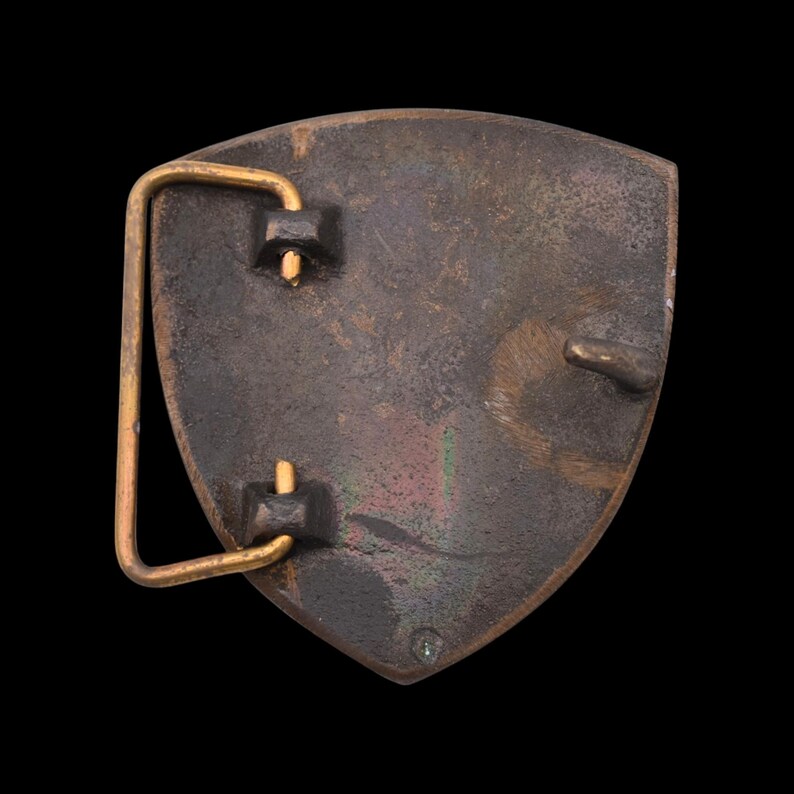 May include: A vintage belt buckle with a shield shape. The buckle is made of a dark, aged metal with a gold-colored bar and attachment points. The surface shows signs of wear and patina, suggesting age.