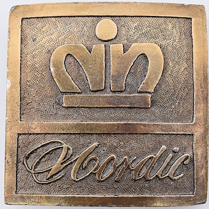 K&ouml;nnte beinhalten: Eine goldfarbene Metall-G&uuml;rtelschnalle mit einer stilisierten Krone und dem Wort "Nordic" darunter.