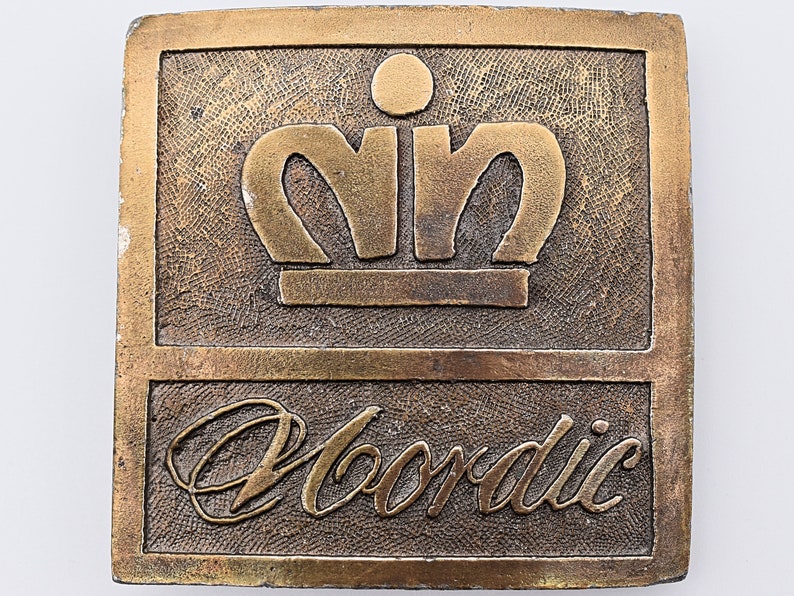 K&ouml;nnte beinhalten: Eine goldfarbene Metall-G&uuml;rtelschnalle mit einer stilisierten Krone und dem Wort "Nordic" darunter.